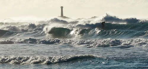 This landscape photograph, taken in the afternoon during the autumn season, depicts a surfer riding heavy waves off the coast of Sennen Cove in the United Kingdom. The sea is turbulent, with large waves dominating the scene, showcasing the raw power of nature. In the background, the iconic Longships Lighthouse stands tall on the horizon, marking a well-known landmark off the Cornish coast. The image captures the dynamic interaction between the surfer and the relentless waves, set against the dramatic backdrop of the open sea. The scene is quintessential of the United Kingdom's rugged coastline, with the Longships Lighthouse providing a focal point that highlights the maritime heritage of the area. The combination of the powerful waves, the lighthouse, and the coastal landscape emphasizes the natural beauty and challenging conditions that make this area popular for surfing.